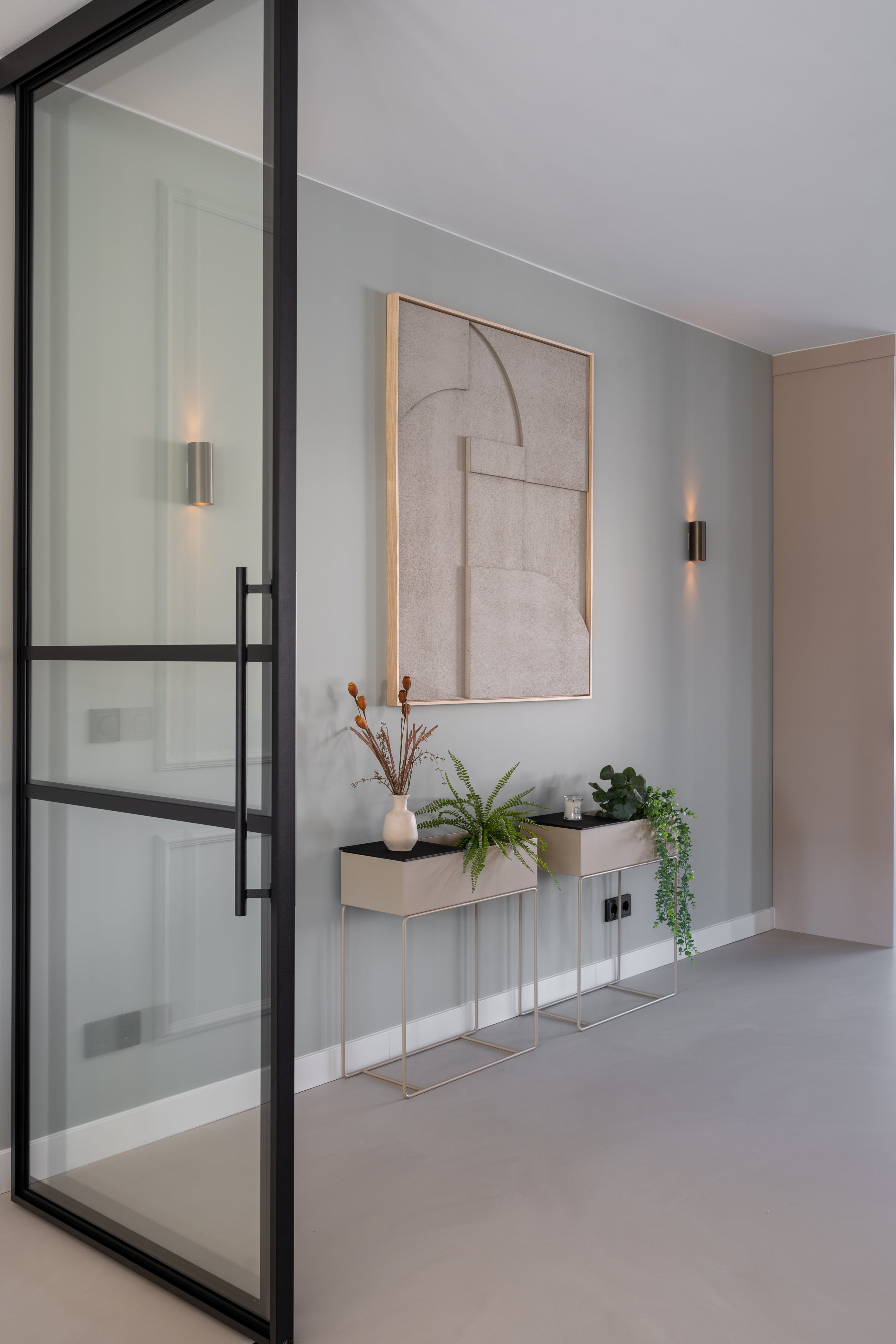 Minimalistische hal met glazen deur, kunst aan de muur en twee smalle tafels met planten.