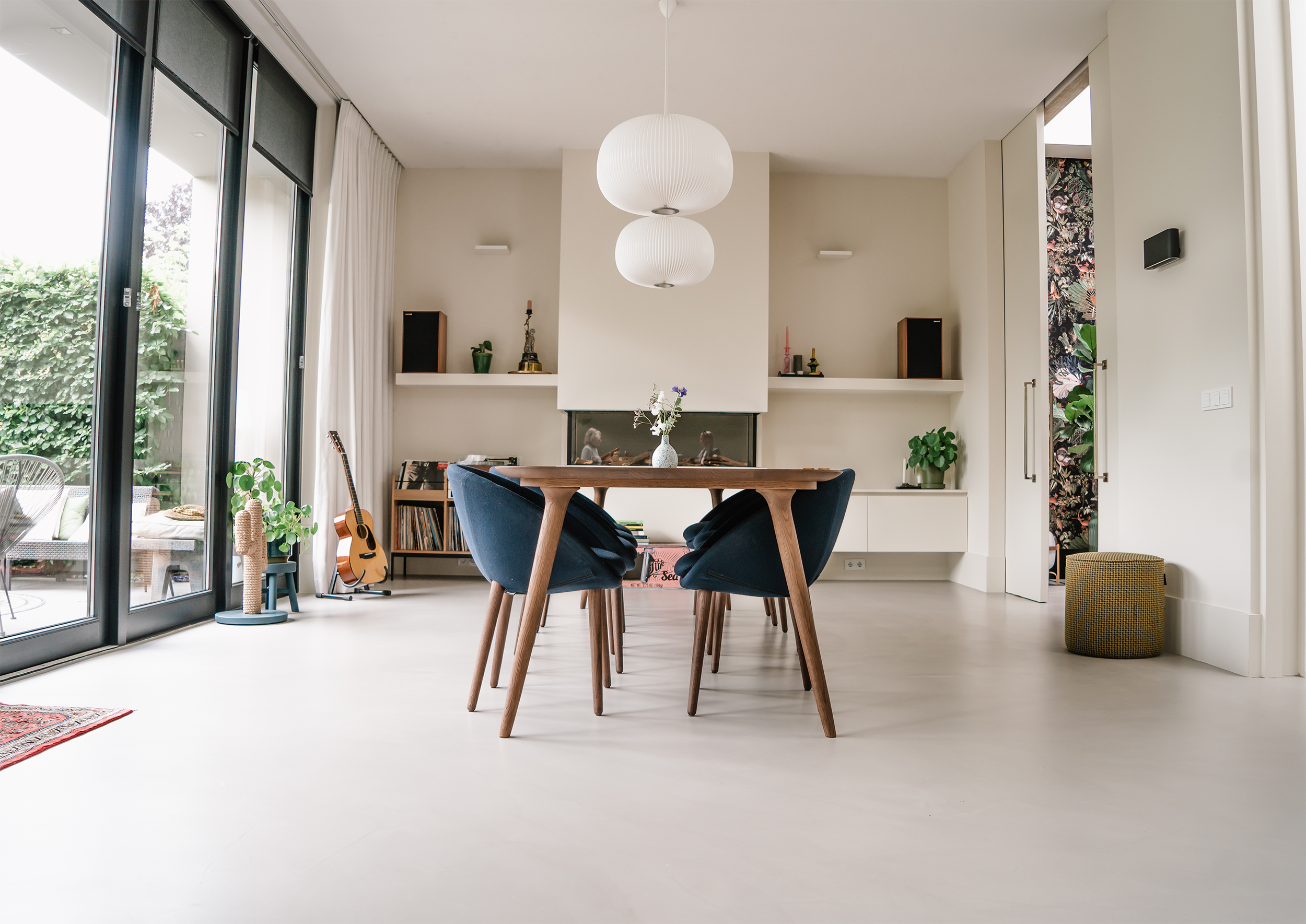 Lichte eetkamer met houten tafel, blauwe stoelen en grote ramen naar de tuin.