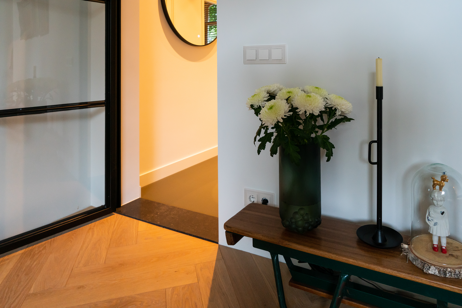 Donkere houten vloer met groen glazen vaas met bloemen, zwarte kandelaar en wanddecoratie in een hal.