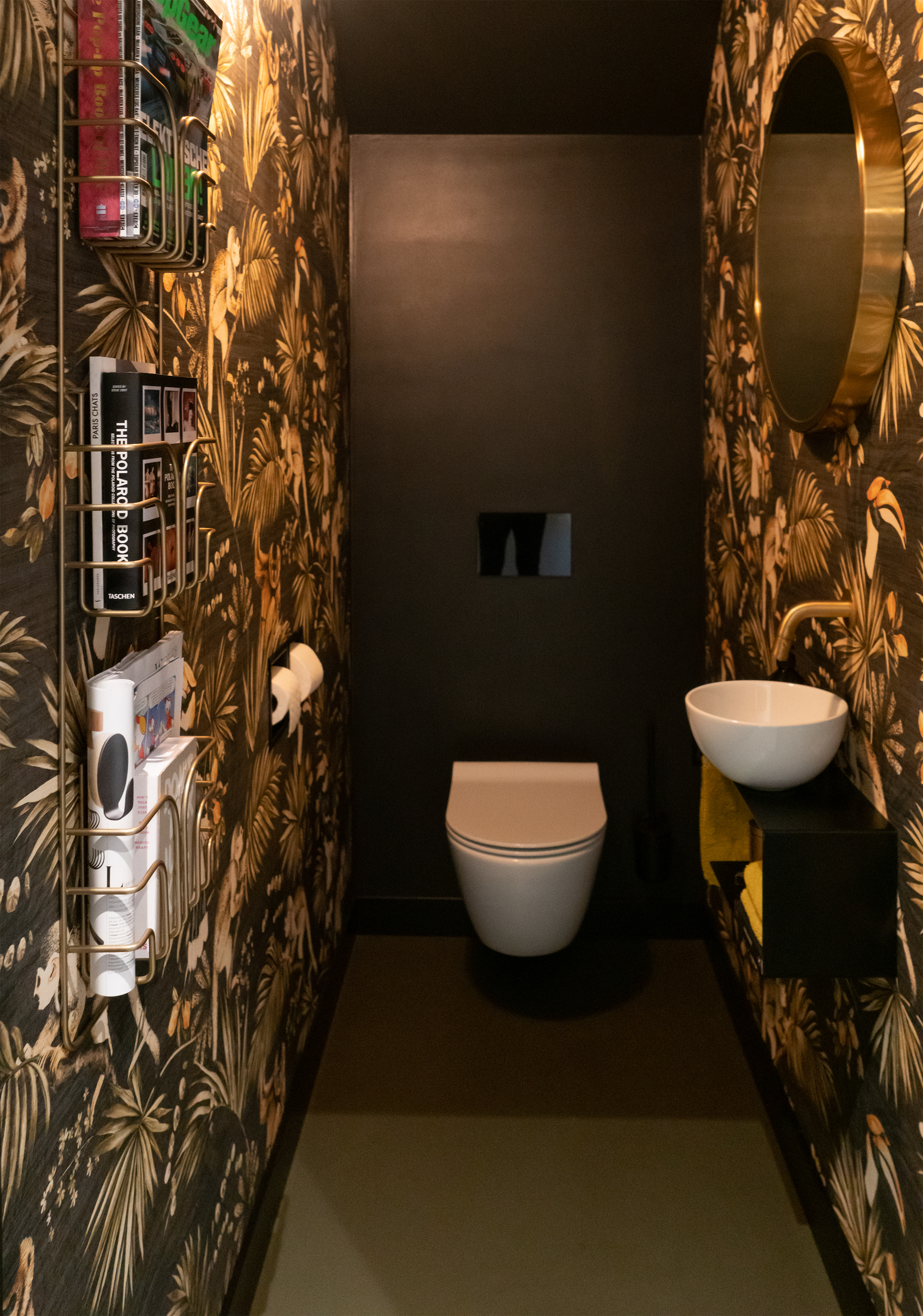 Smalle toiletruimte met donkere botanische behang, ronde spiegel en gouden accenten.