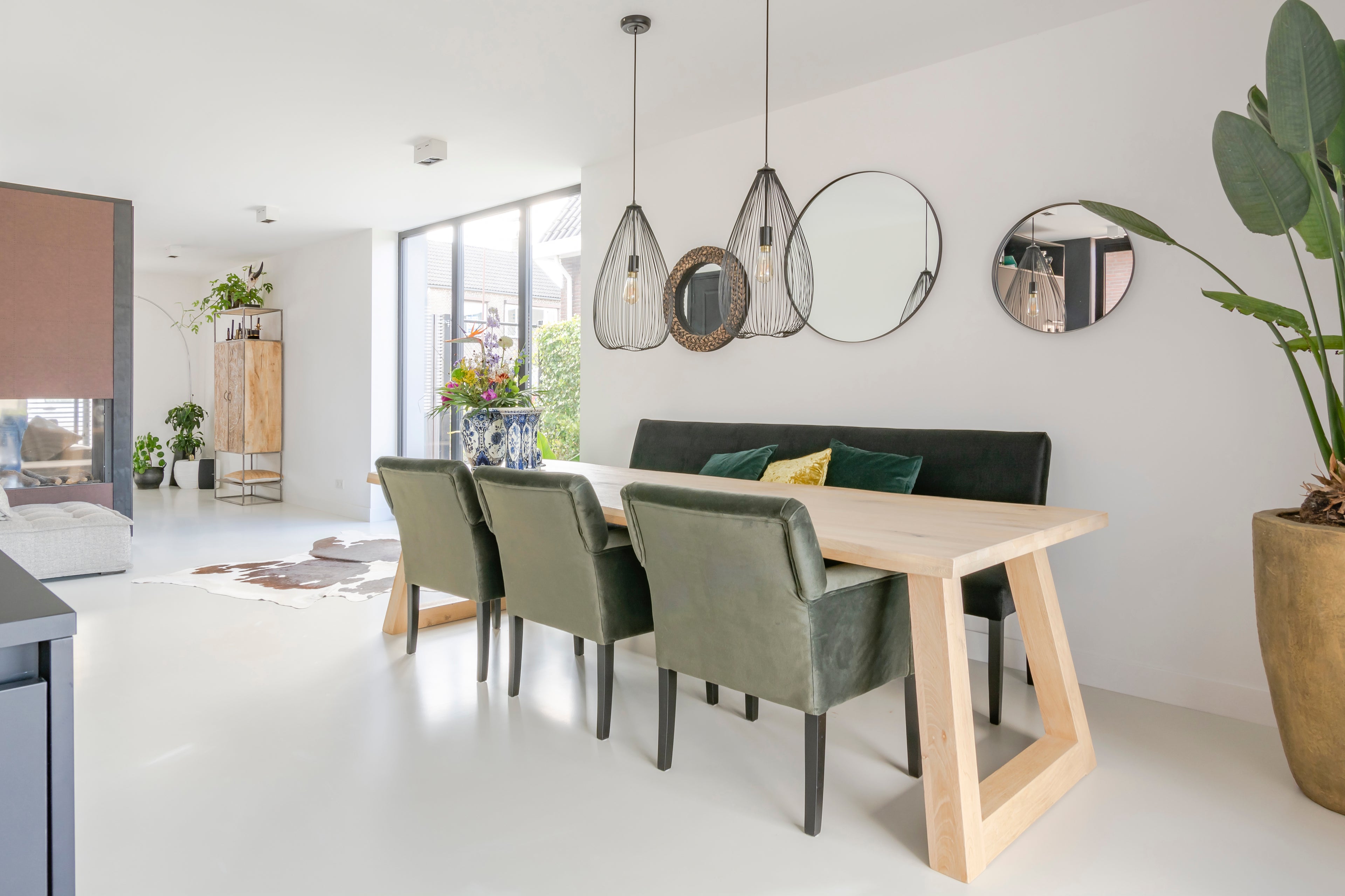 Lichte eetruimte met houten tafel, groene stoelen, hanglampen en decoratieve ronde spiegels.