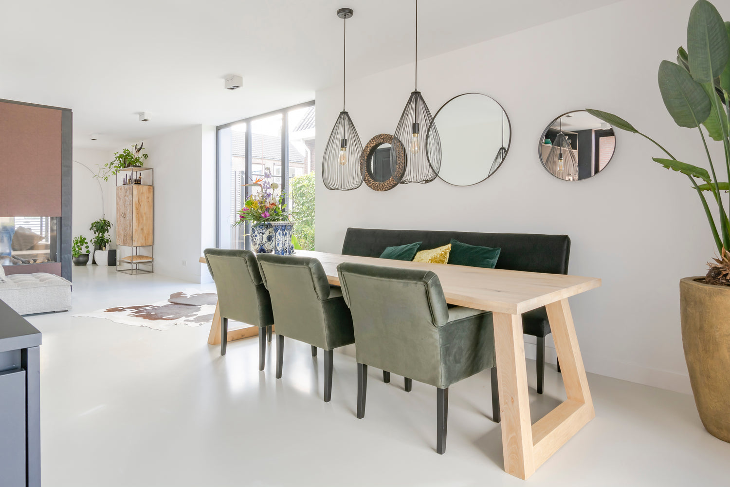 Lichte eetkamer met houten tafel, groene stoelen, hanglampen en ronde spiegels aan de muur.