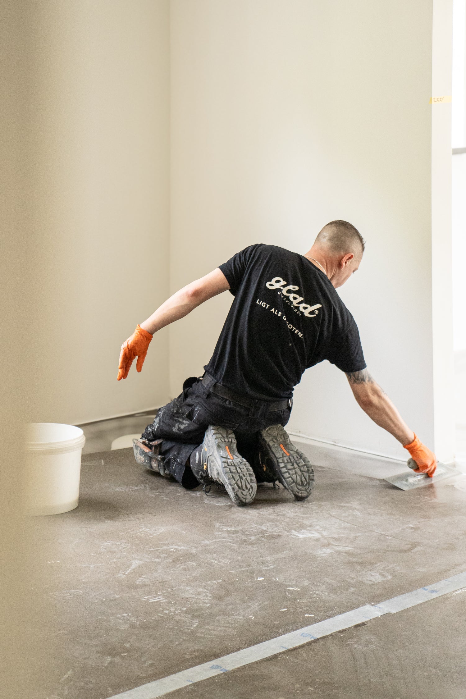 Vloerspecialist van Glad Gietvloeren brengt gietvloer aan op betonvloer in lege ruimte, geknield met troffel en handschoenen.