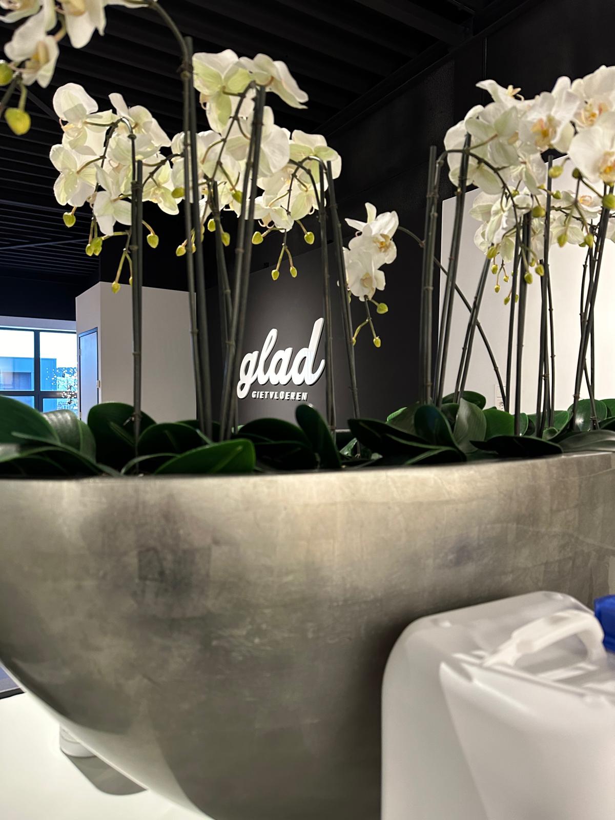 Grote zilverkleurige plantenbak met witte orchideeën en het logo ‘glad’ op zwarte wand.