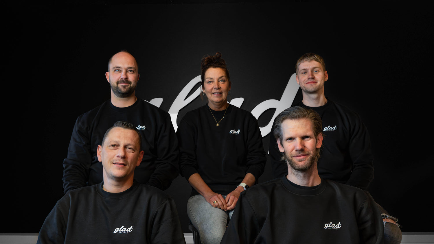 Teamfoto van de werknemers van Glad Gietvloeren, iedereen draagt zwarte trui met het logo van Glad. Zwarte achtergrond met het logo van Glad.