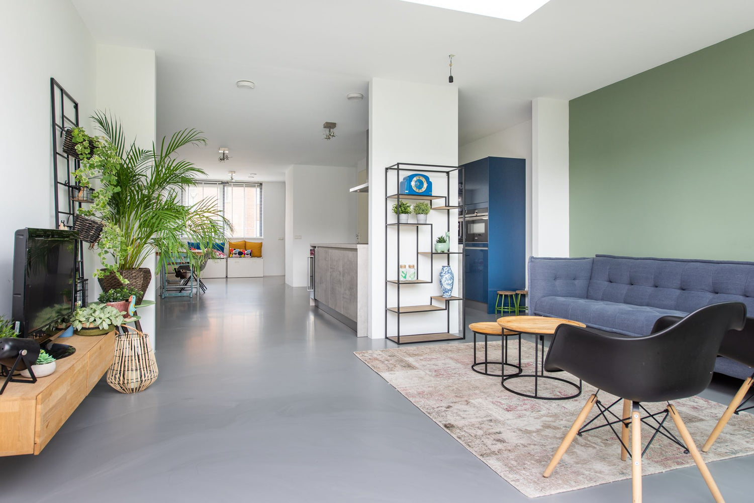 Moderne woonkamer met groene accentmuur, blauwe bank, zwarte stoel en veel kamerplanten.