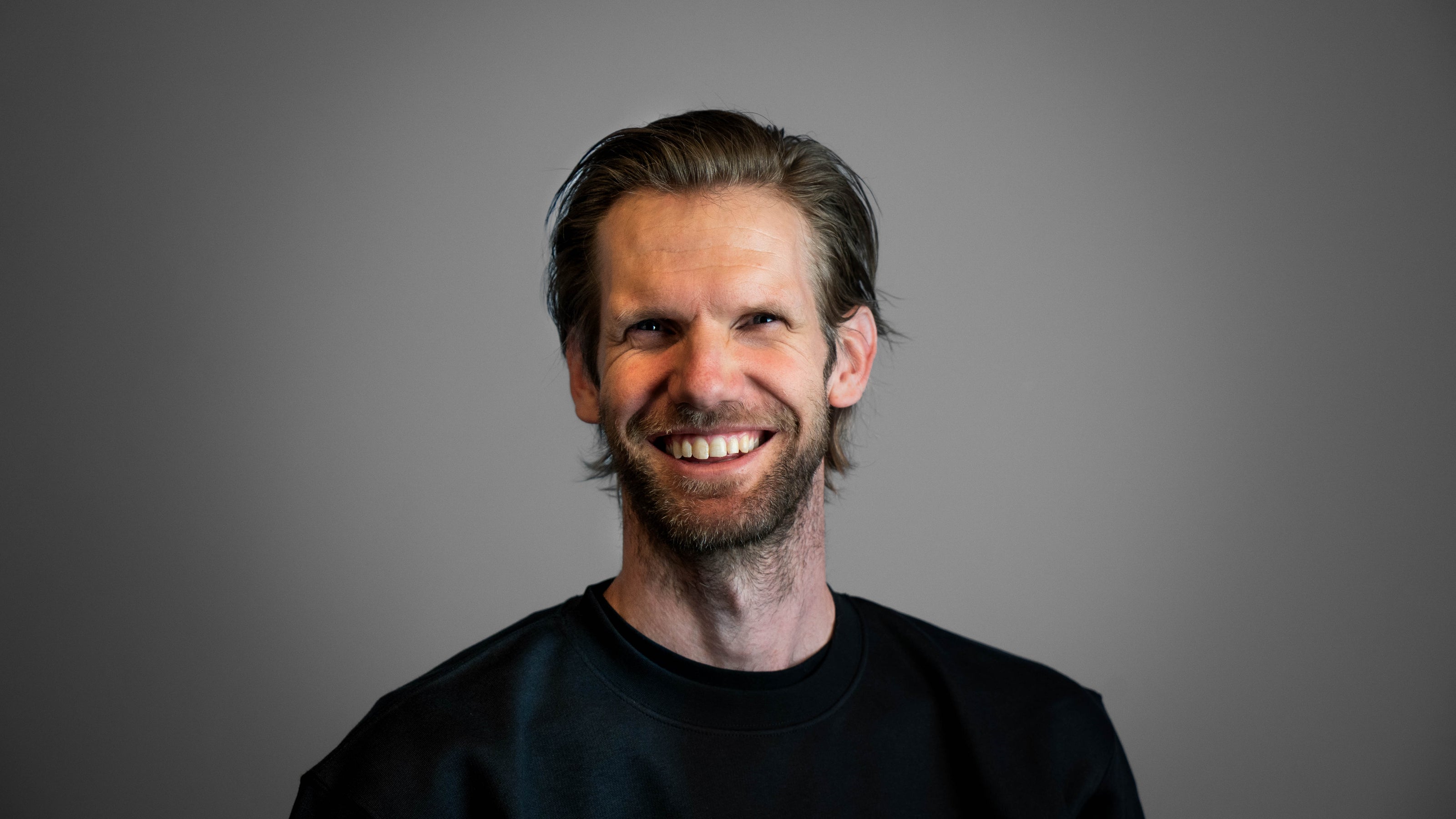 Lachende man met korte baard, zwarte trui op een grijze achtergrond. Medewerker bij glad gietvloeren.