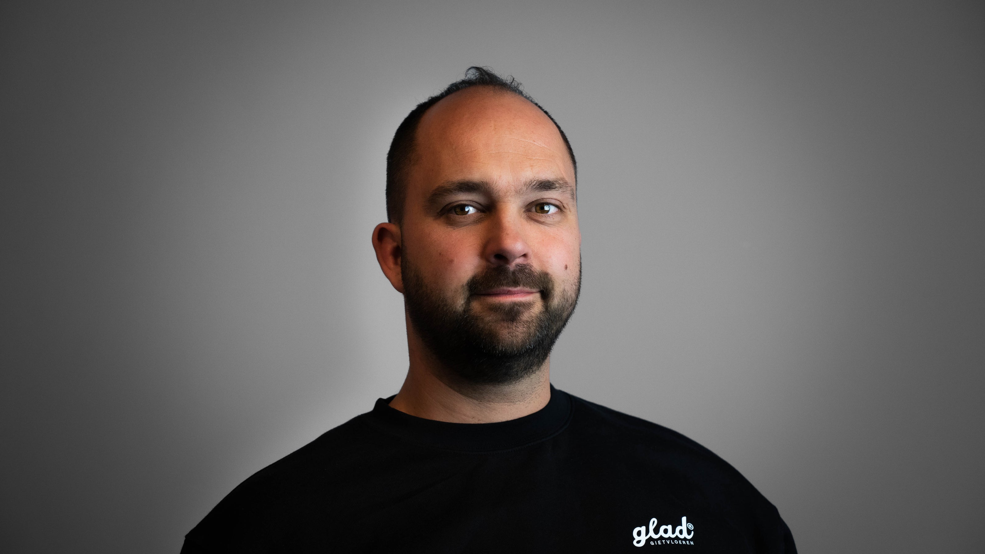 Vriendelijke man met korte baard, glimlach, zwarte trui met logo van Glad Gietvloeren. Grijze achtergrond. Eigenaar van Glad Gietvloeren.