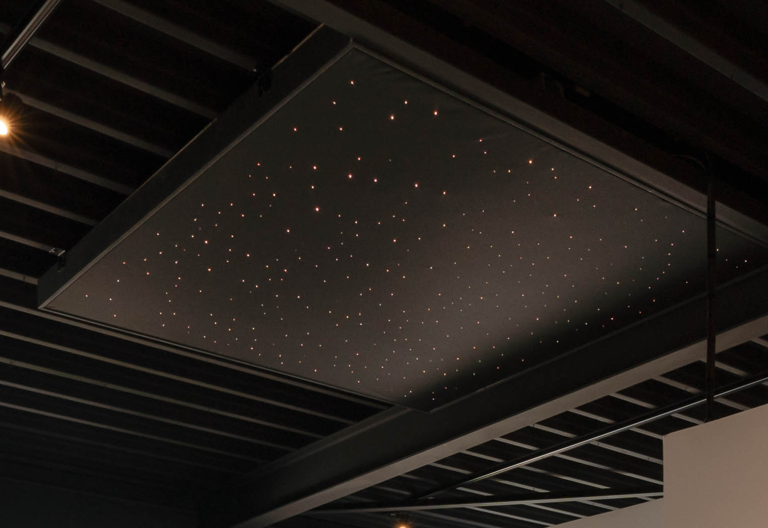 Zwart plafondpaneel met subtiele sterrenhemelverlichting in een ruimte met industriële plafondbalken.